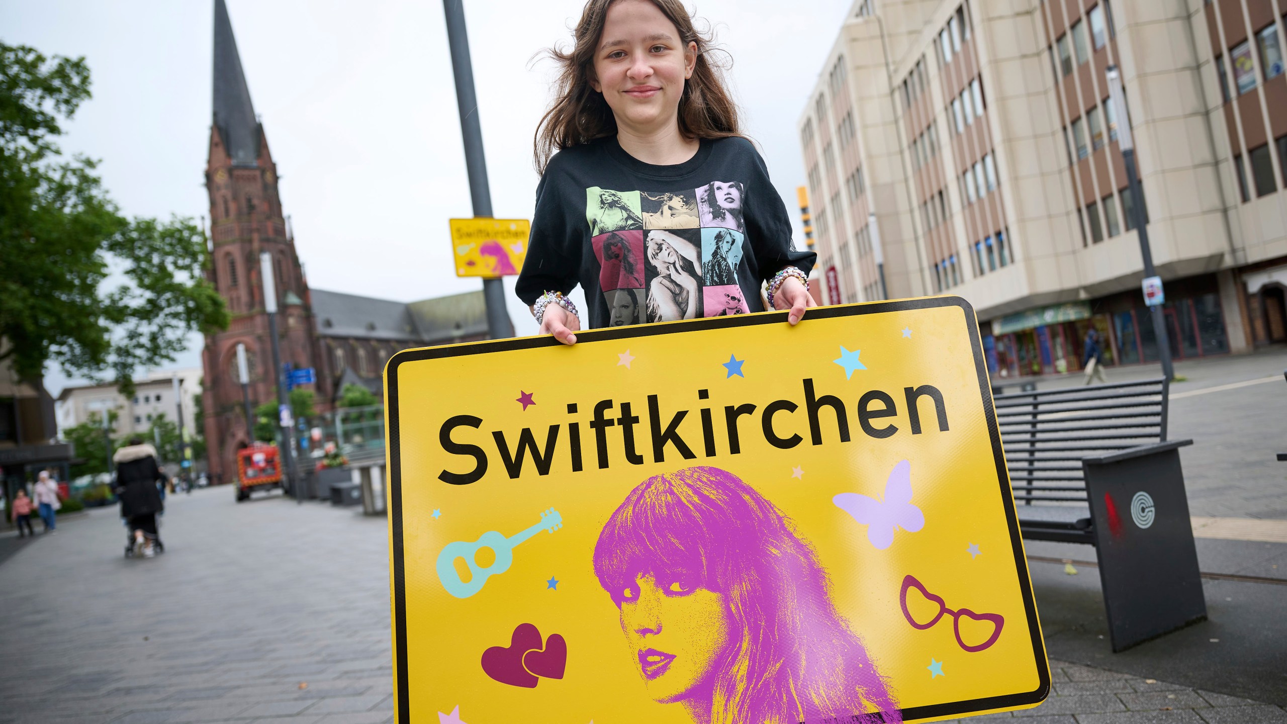 Schoolgirl Aleshanee Westhoff shows a "Swiftkirchen" town sign in honor of musician Taylor Swift in Gelsenkirchen, Germany, Tuesday, July 2, 2024. The Swifties are about to take over the German city of Gelsenkirchen, where American superstar Taylor Swift is set to give three concerts of her Eras Tour later this month. In honor of the singer, the city renamed itself “Swiftkirchen" — at least temporarily to welcome the tens of thousands of fans who are expected to come for the Eras Tour shows on July 17, 18 and 19, German news agency dpa reported. (Bernd Thissen/dpa via AP)