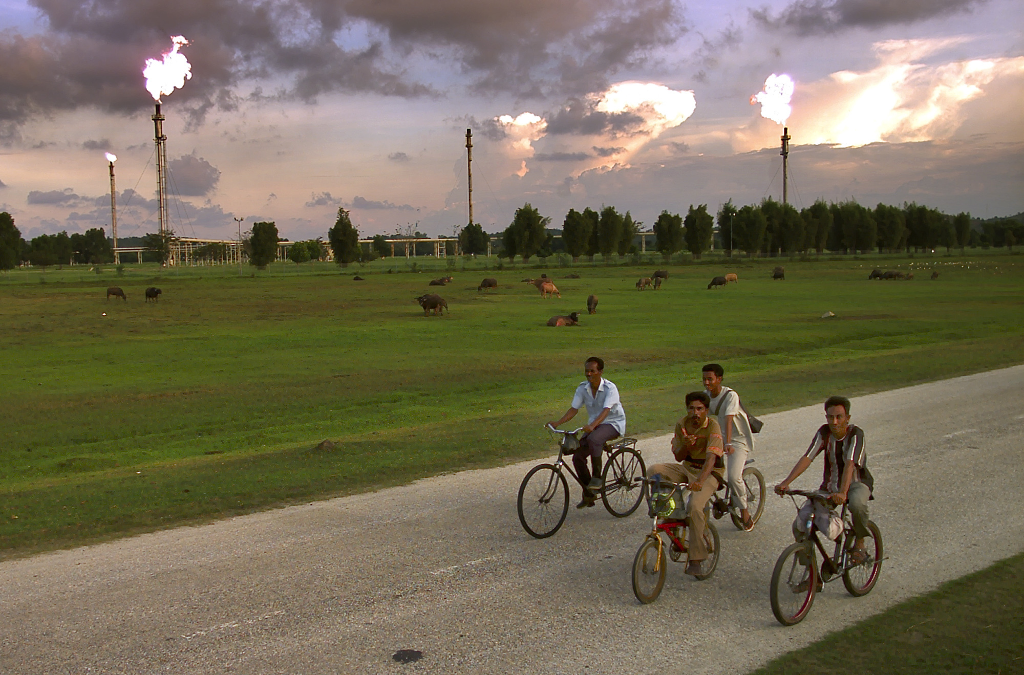 FILE- Acehnese men bicycle to work past an Exxon Mobil gas production facility in Lhokseumawe, Aceh province, Indonesia, April 4, 2001.(AP Photo/Ed Wray, File)