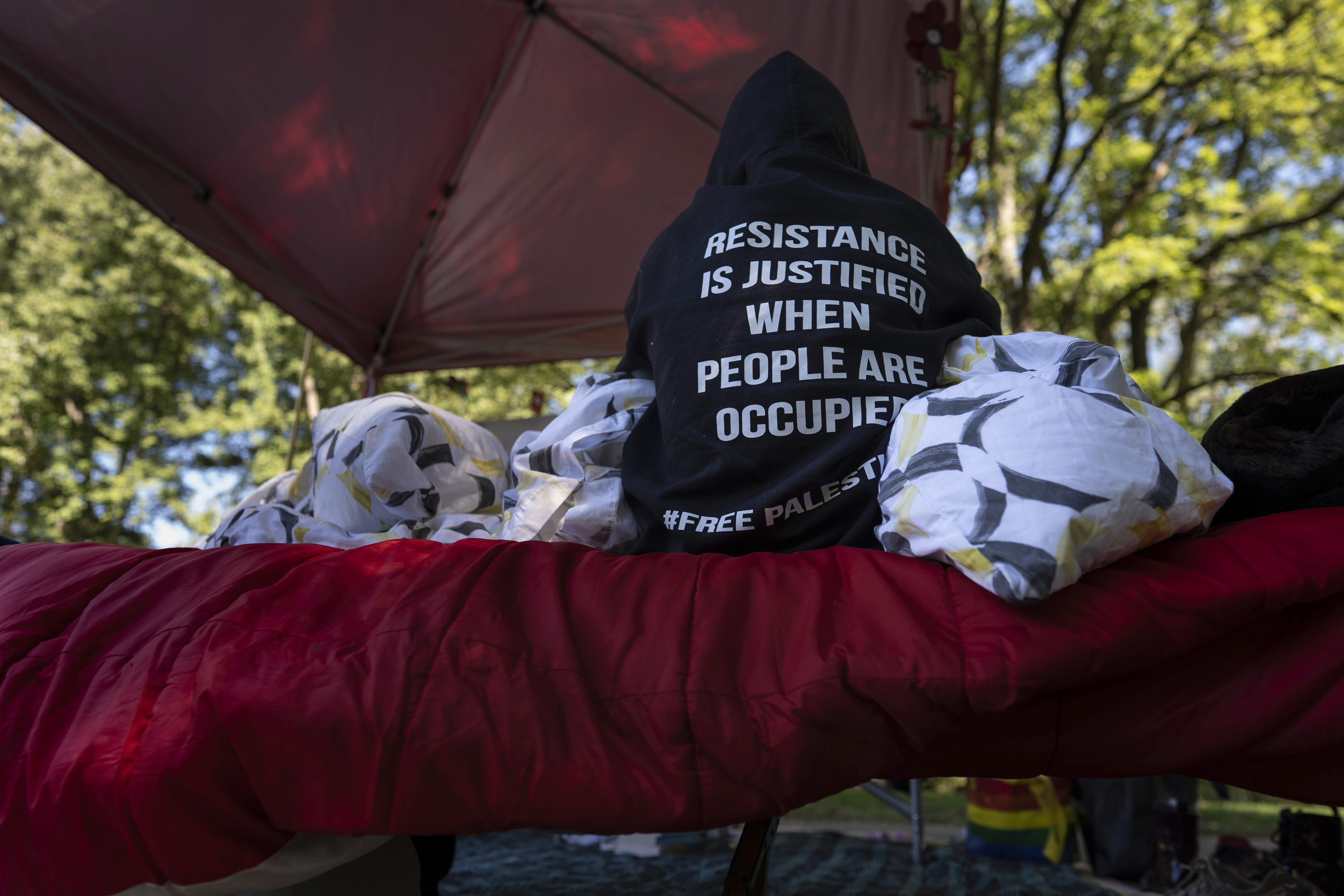 A protester sits on a cot on the side of the road near the driveway of Ohio Democratic Rep. Greg Landsman's Cincinnati residence wearing a sweatshirt that reads "Resistance is justified when people are occupied #Free Palestine," Monday, Oct. 7, 2024. (AP Photo/Carolyn Kaster)