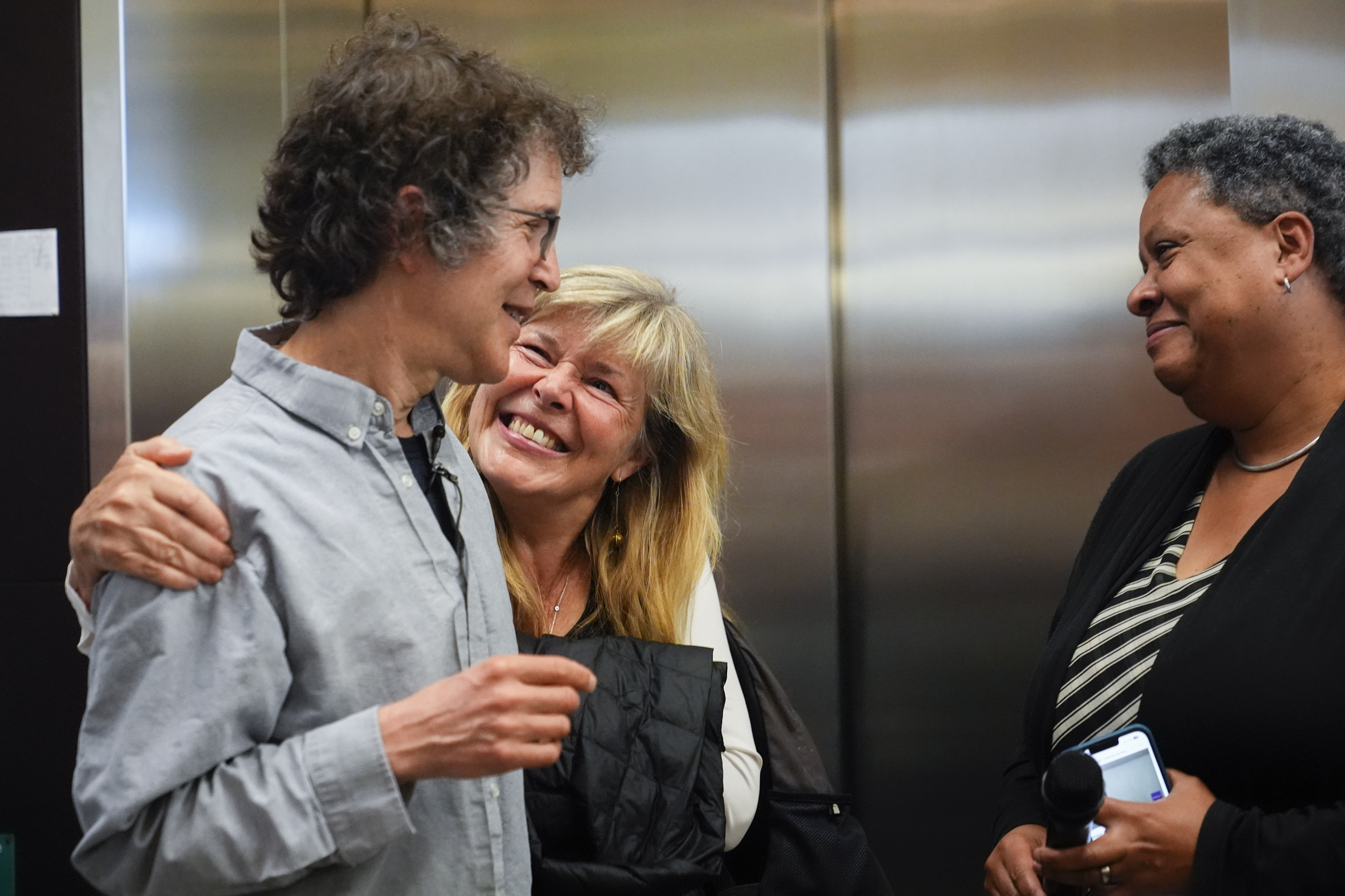 Hannele Ruohola-Baker hugs her husband American biochemist David Baker, 2024 Nobel Prize winner in Chemistry, at the University of Washington on Wednesday, Oct. 9, 2024, in Seattle. Lynda Stuart, executive director at the Institute for Protein Design, looks on at right. (AP Photo/Lindsey Wasson)