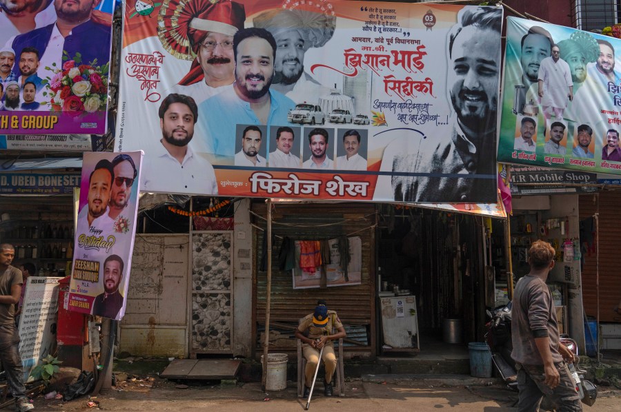 A police person sits below a large hoarding showing a portrait in red turban of Baba Siddique, a senior politician, at the spot where Siddique was shot at outside his son's office in Mumbai and later succumbed to his wounds in a hospital on Saturday night, in Mumbai, India, Sunday, Oct. 13, 2024. (AP Photo/Rafiq Maqbool)