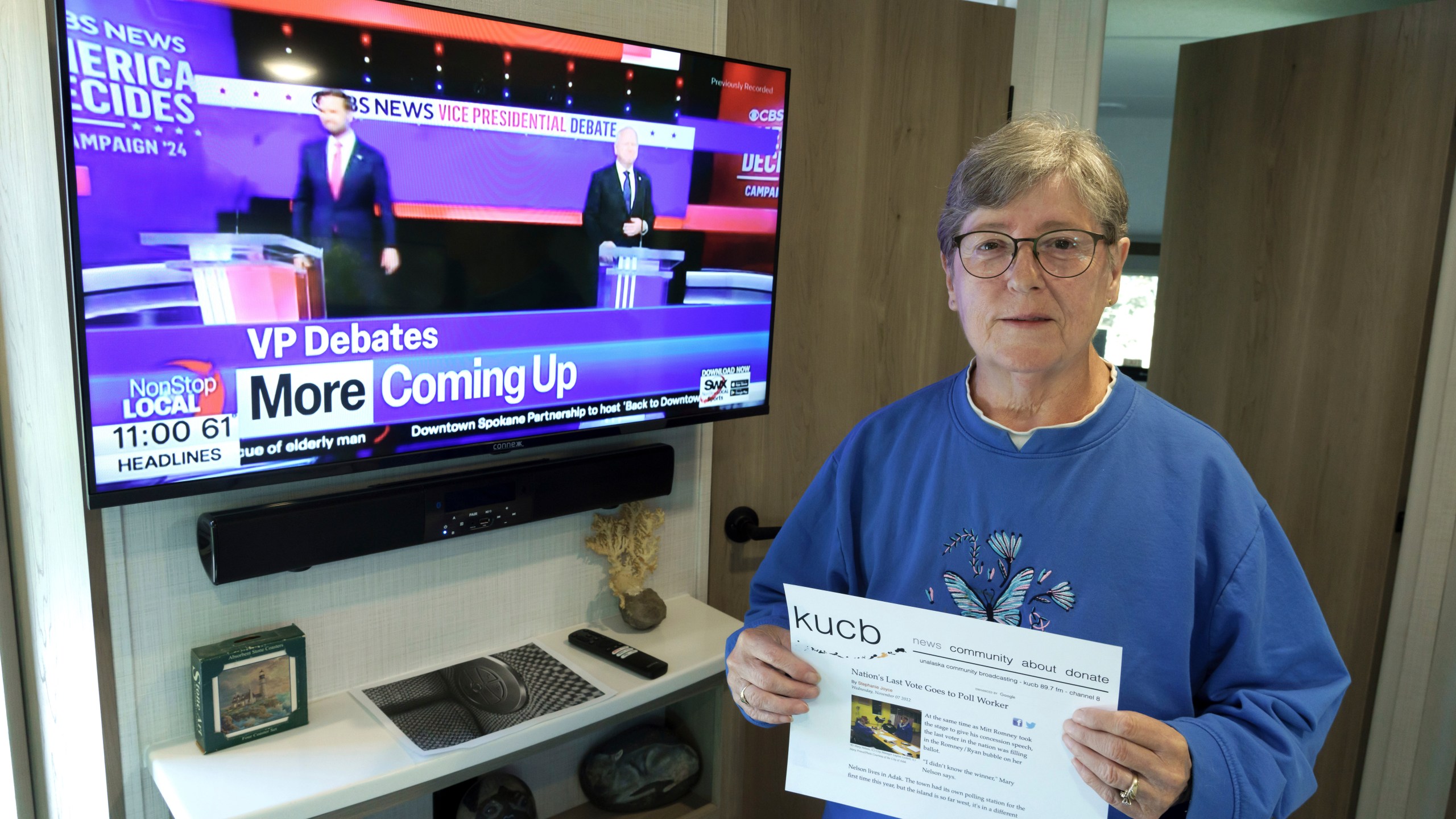 Mary Nelson, of Mead, Wash., poses for a photo at her home on Wednesday, Oct. 2, 2024, as a local TV station broadcasts a news story about the vice presidential debate. Nelson is holding a printout of a radio station website news story that says she is thought to be the last person in the U.S. to cast a vote in the 2012 presidential election when she voted for Mitt Romney just before the polls closed in the remote village of Adak, Alaska. (AP Photo/Ted S. Warren)