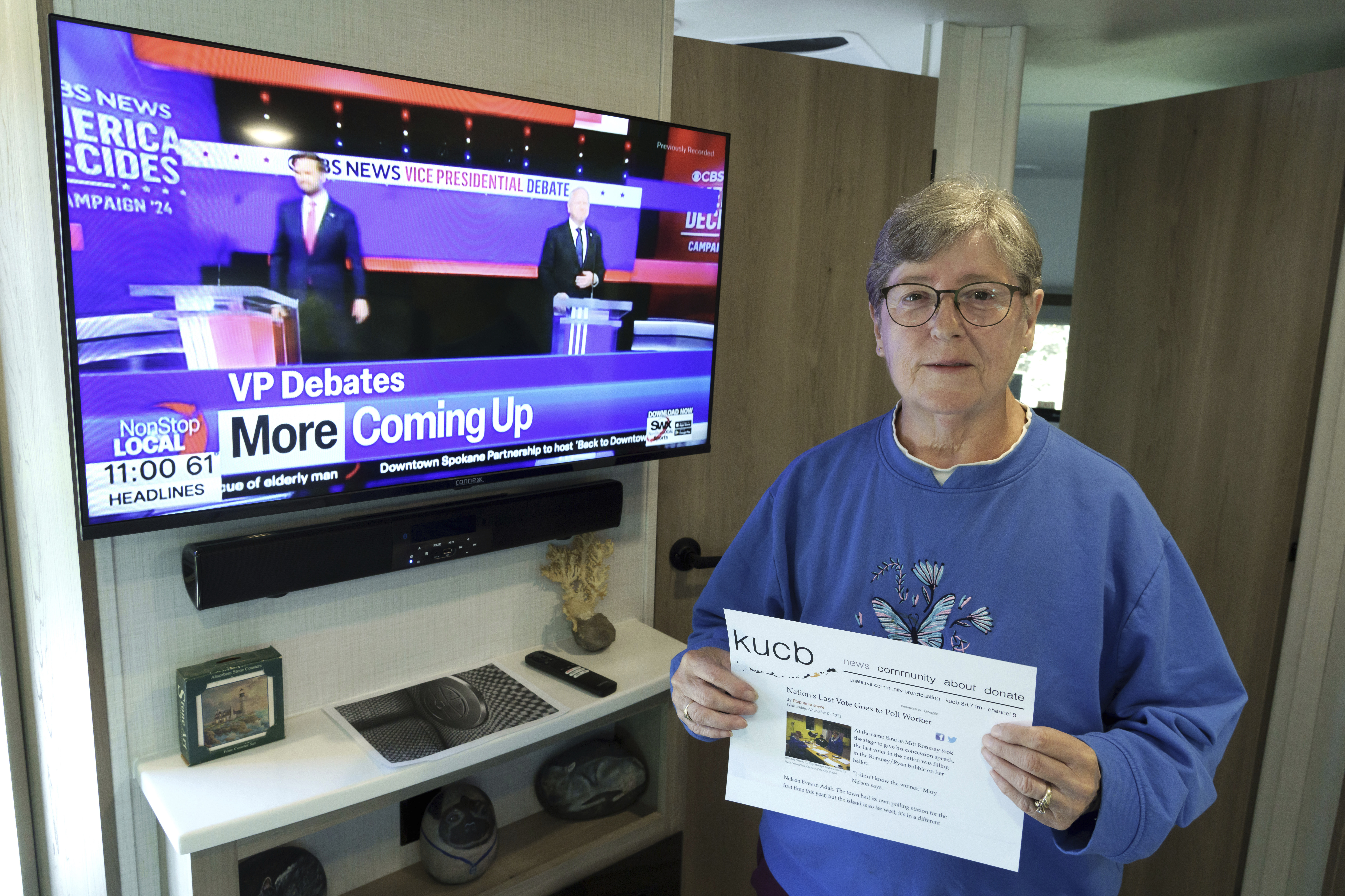 Mary Nelson, of Mead, Wash., poses for a photo at her home on Wednesday, Oct. 2, 2024, as a local TV station broadcasts a news story about the vice presidential debate. Nelson is holding a printout of a radio station website news story that says she is thought to be the last person in the U.S. to cast a vote in the 2012 presidential election when she voted for Mitt Romney just before the polls closed in the remote village of Adak, Alaska. (AP Photo/Ted S. Warren)