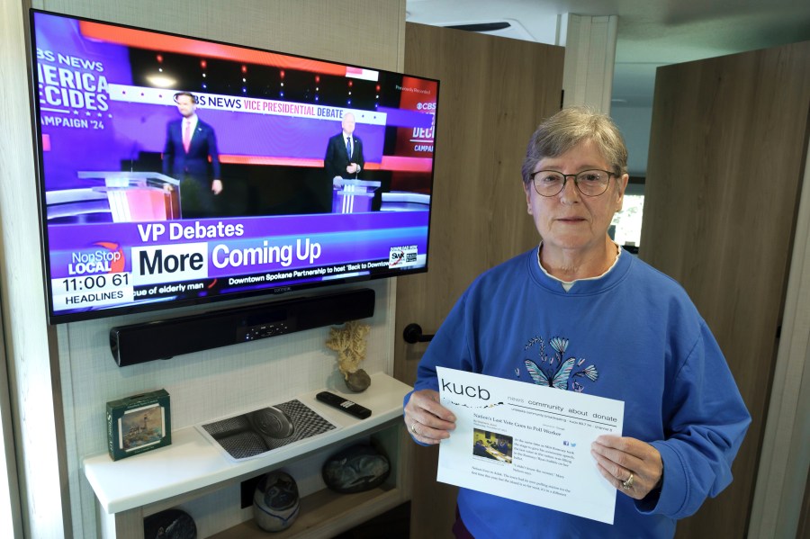 Mary Nelson, of Mead, Wash., poses for a photo at her home on Wednesday, Oct. 2, 2024, as a local TV station broadcasts a news story about the vice presidential debate. Nelson is holding a printout of a radio station website news story that says she is thought to be the last person in the U.S. to cast a vote in the 2012 presidential election when she voted for Mitt Romney just before the polls closed in the remote village of Adak, Alaska. (AP Photo/Ted S. Warren)