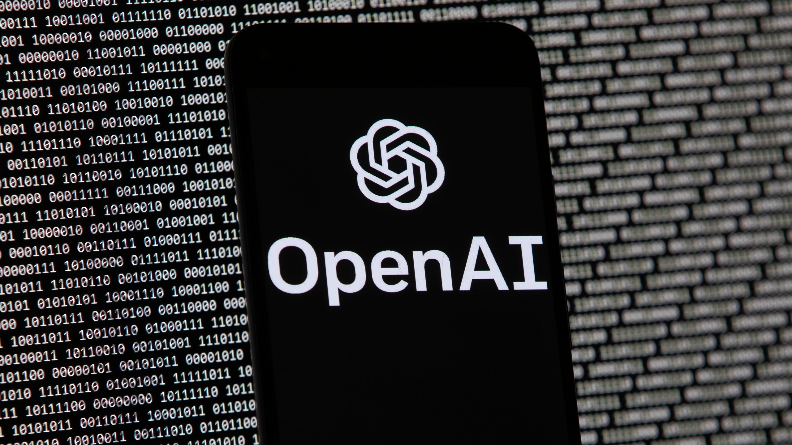 FILE - The OpenAI logo appears on a mobile phone in front of a computer screen with random binary data, March 9, 2023, in Boston. (AP Photo/Michael Dwyer, File)