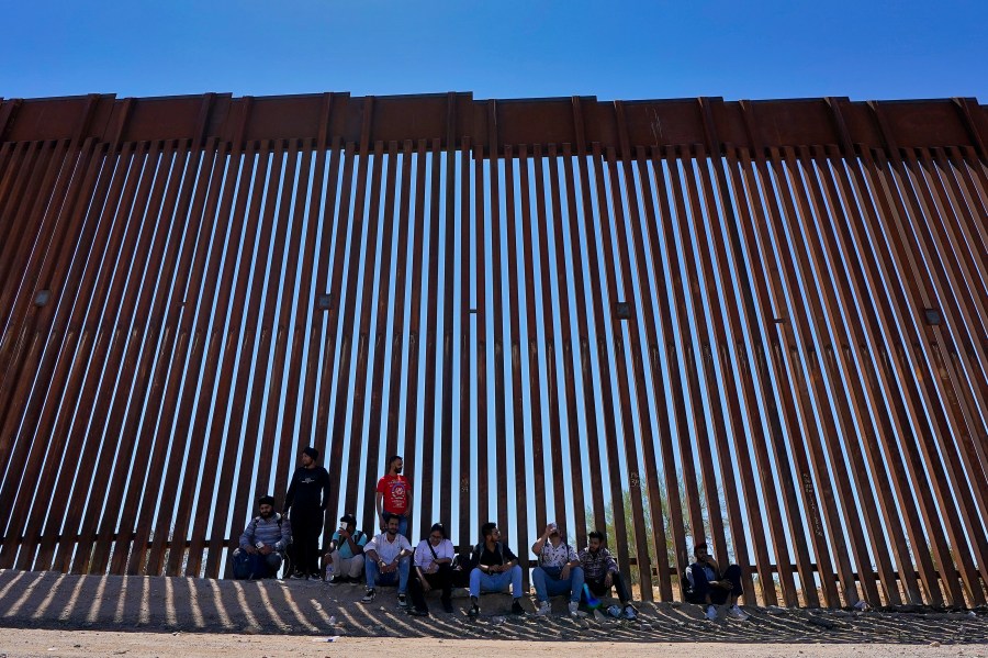 FILE - A group claiming to be from India sit in the shade of the border wall as they wait to be picked up by Border Patrol after crossing through the border fence in the Tucson Sector of the U.S.-Mexico border, Aug. 29, 2023, in Organ Pipe Cactus National Monument near Lukeville, Ariz. (AP Photo/Matt York, File)