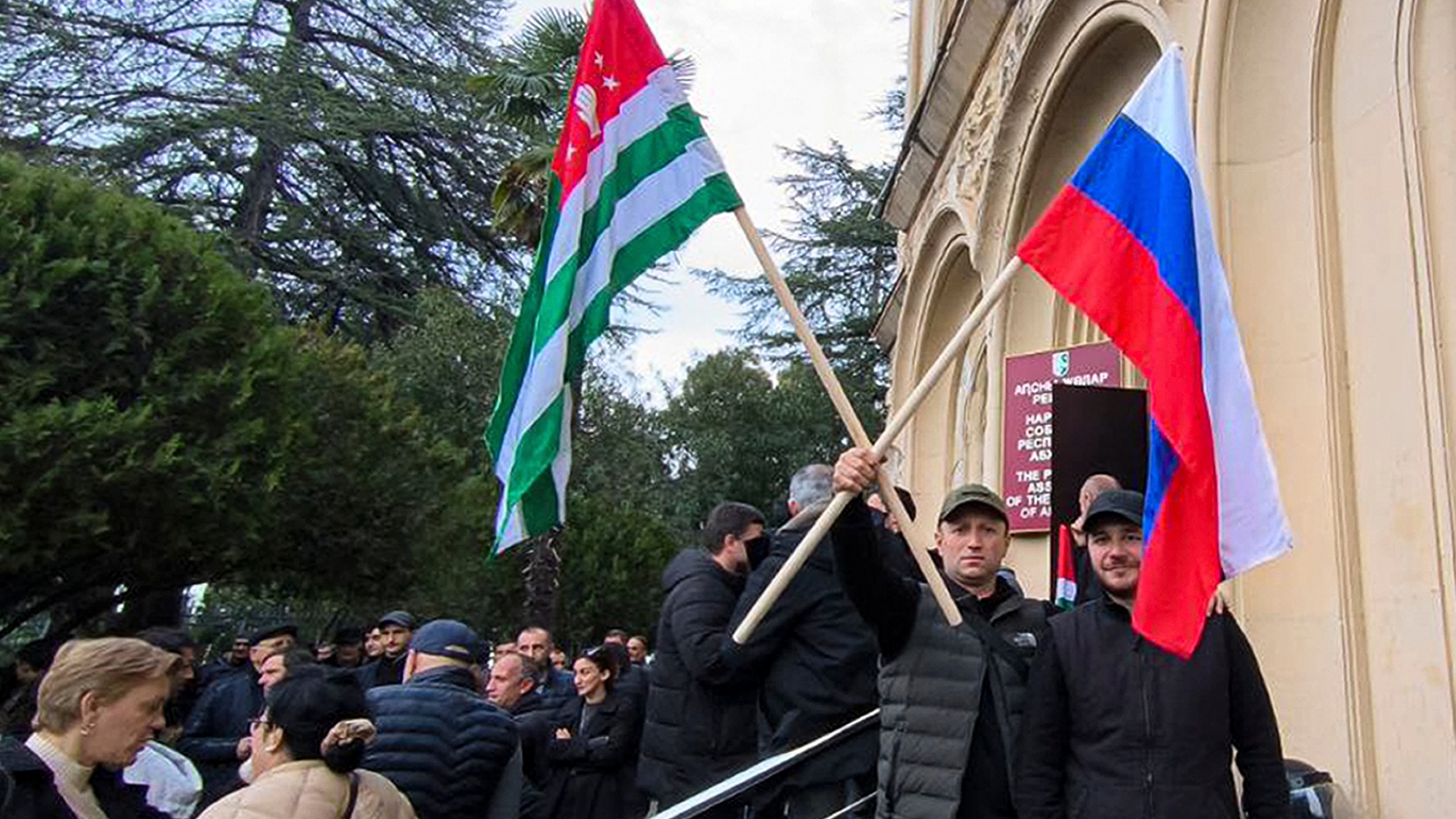 In this photo taken from video released by AIASHARA Independent Agency, Protesters, two of them hold an Abkhazian and Russian flags, gather outside the parliament building of the Georgian separatist region of Abkhazia as tensions flared over a proposed pact that would allow Russians to buy apartments in the region, Georgia, on Friday, Nov. 15, 2024, (AIASHARA Independent Agency via AP)