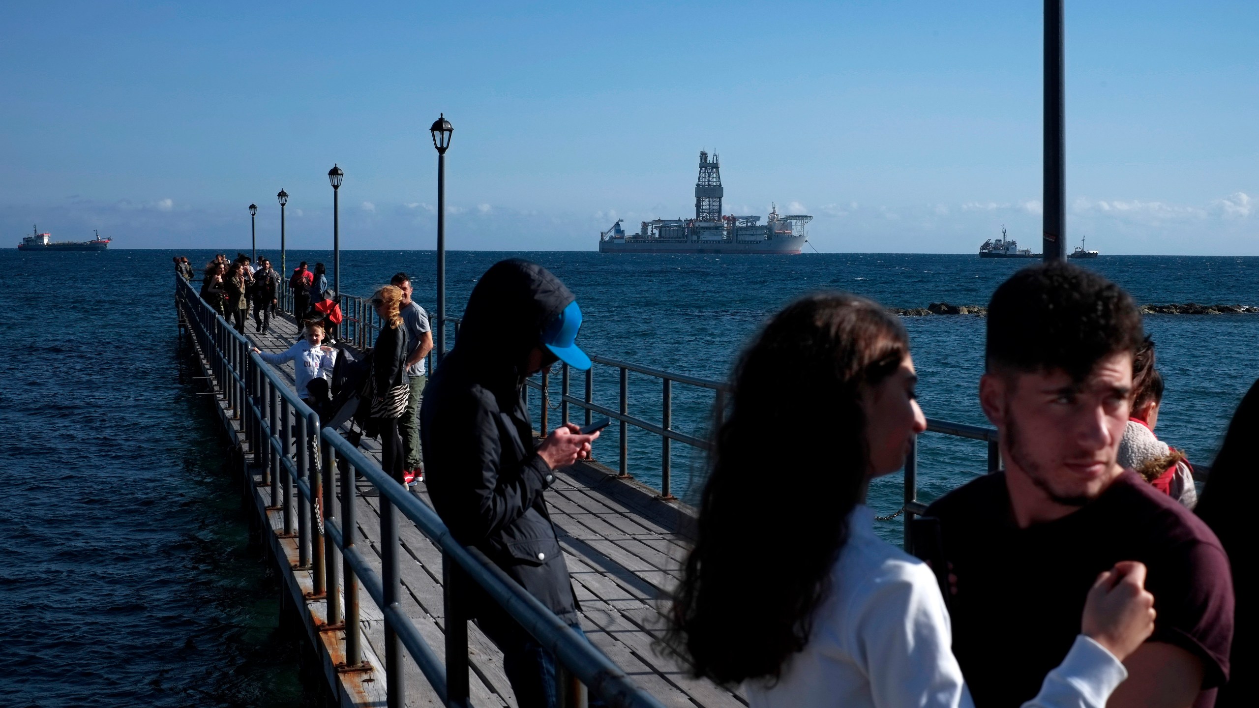 FILE - People walk at a dock with a drilling platform in the background, in the southern coastal city of Limassol, Cyprus, on March 10, 2019. (AP Photo/Petros Karadjias, File)