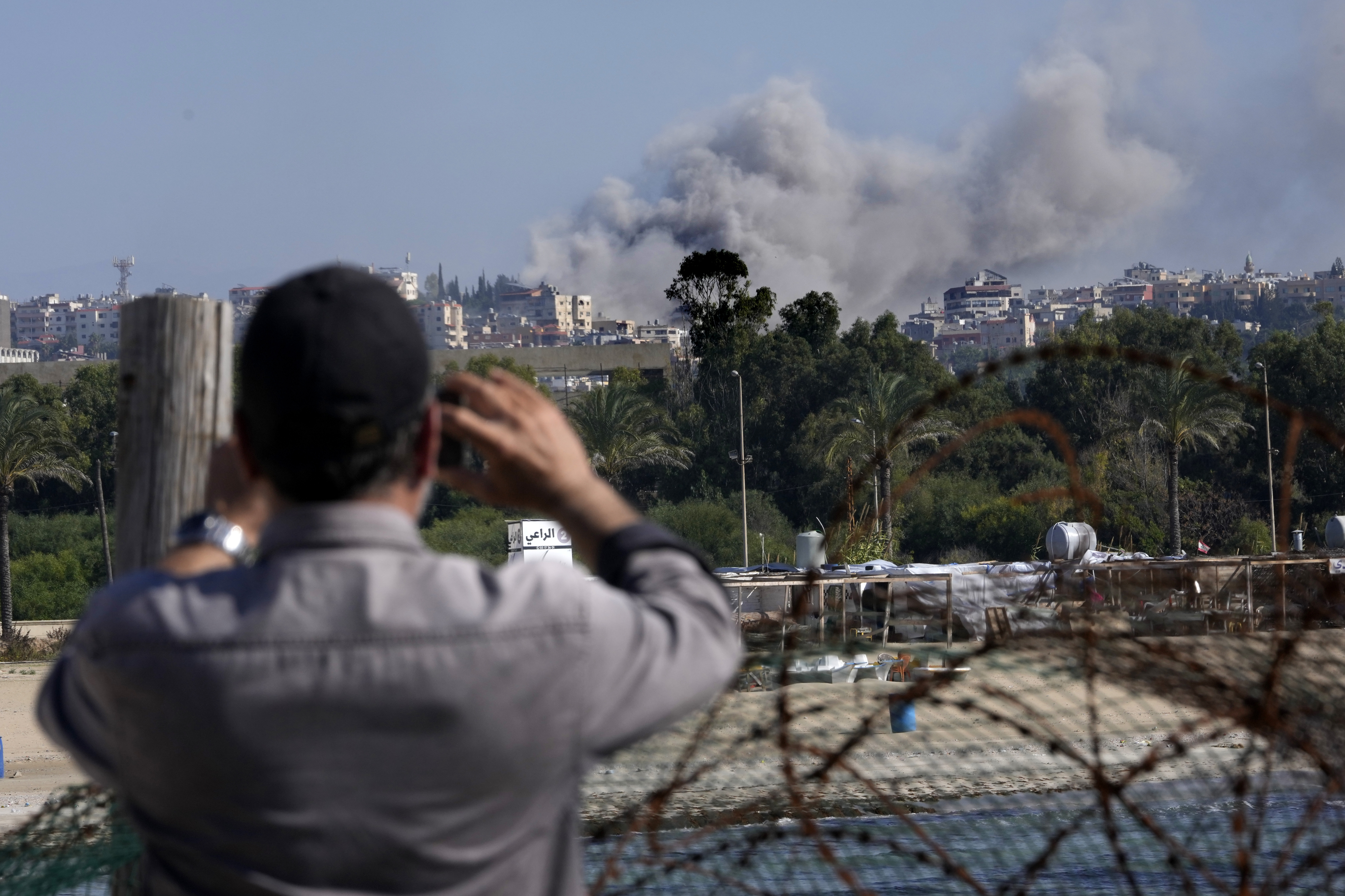 A man takes pictures by his mobile phone for the smoke rises between buildings hit in an Israeli airstrike in Burj al-Shamali village, as it seen from Tyre city, south Lebanon, Friday, Nov. 22, 2024. (AP Photo/Hussein Malla)