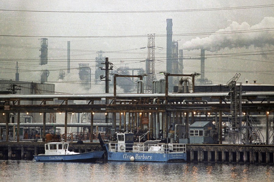 FILE - The Exxon oil refinery stands in Linden, N.J., Jan. 9, 1990, as two clean harbor cleanup boats work in the Arthur Kill waterway. (AP Photo/Mike Derer, File)