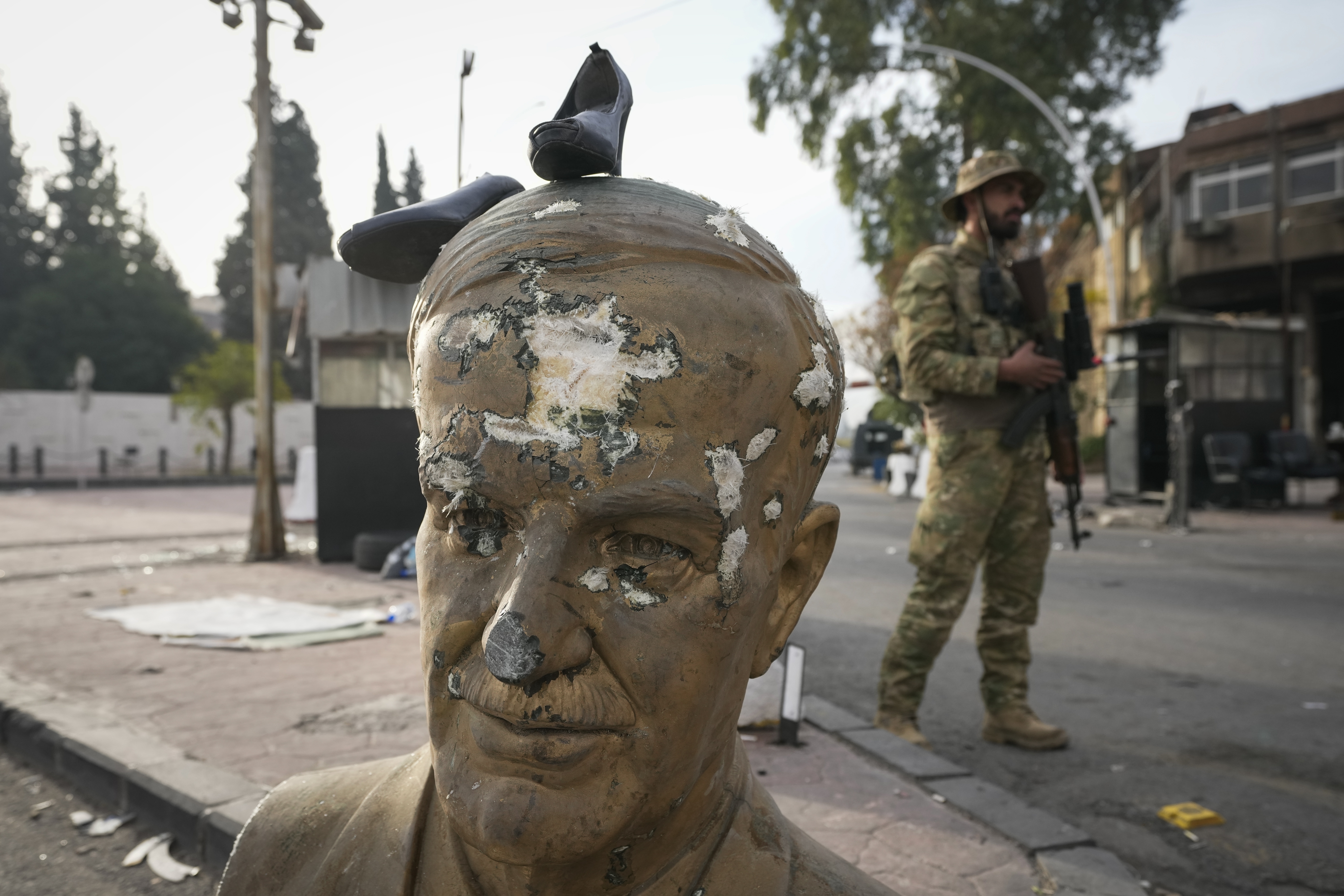 An opposition fighter in military uniform stands next to a broken bust of the late Syrian President Hafez Assad, with a pair of shoes placed on top, at a checkpoint in Damascus, Syria, Wednesday, Dec. 11, 2024. (AP Photo/Hussein Malla)