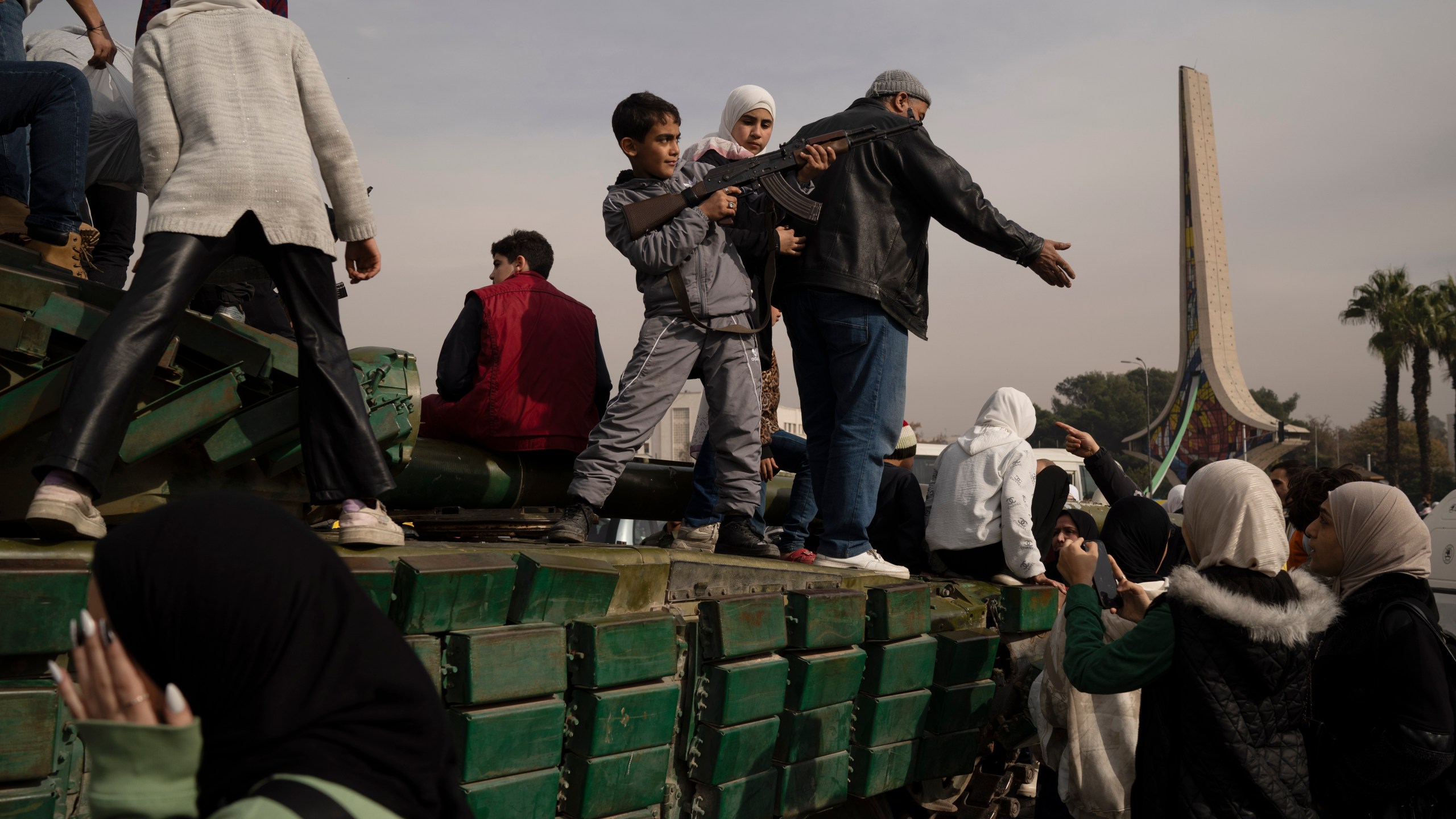 A boy holding a rifle borrowed from a Syrian opposition fighter poses on the top of a government forces tank that was left on a street, at the Umayyad Square in Damascus, Syria, Wednesday, Dec. 11, 2024. (AP Photo/Leo Correa)