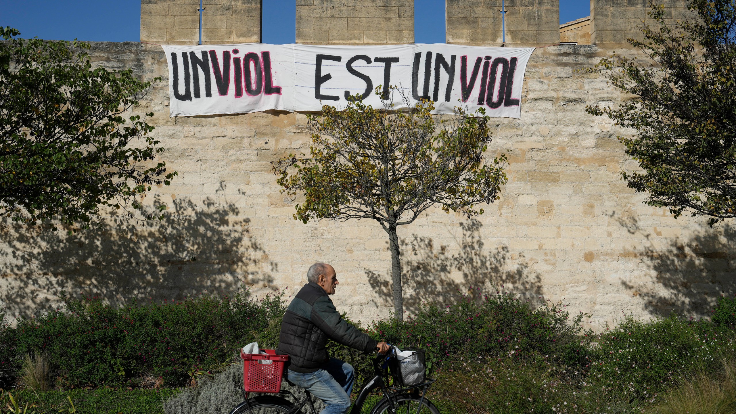 FILE - A man rides a bicycle in front of a banner that reads, "A rape is a rape," in Avignon, southern France, on Oct. 16, 2024. (AP Photo/Lewis Joly, File)