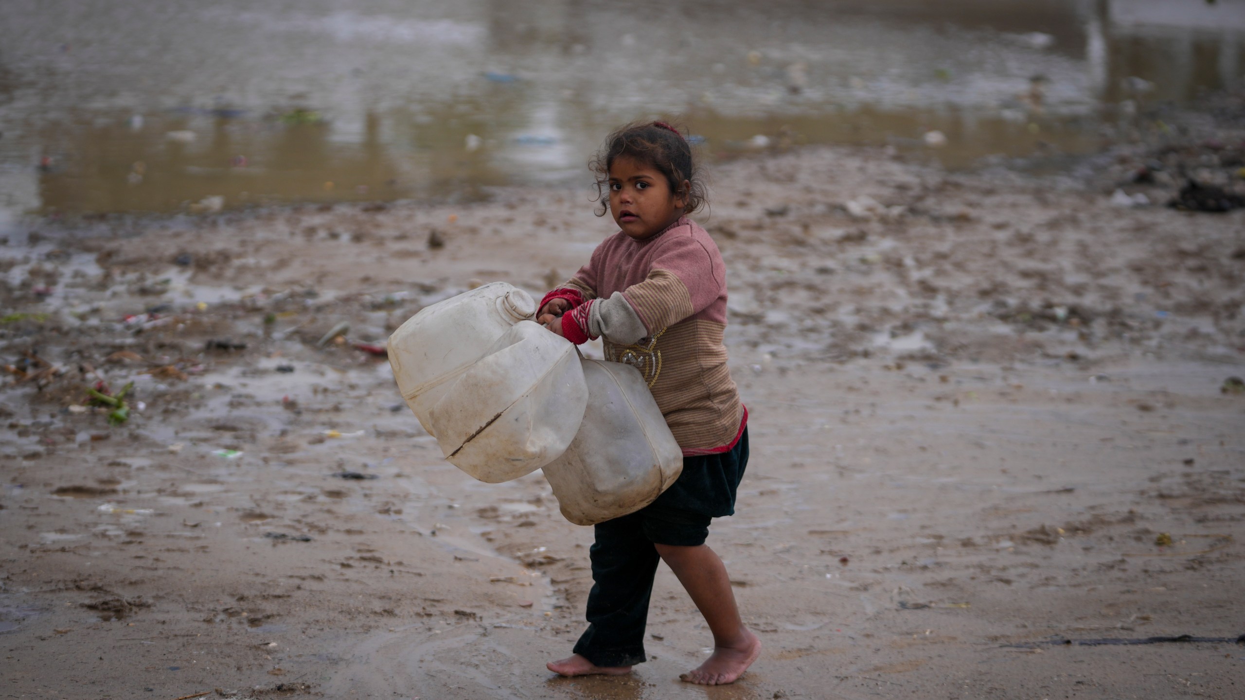 A young girl walks barefoot, carrying empty jerrycans to collect water, after overnight rainfall at the refugee tent camp for displaced Palestinians in Deir al-Balah, central Gaza Strip,, Tuesday, Dec. 31, 2024. (AP Photo/Abdel Kareem Hana)