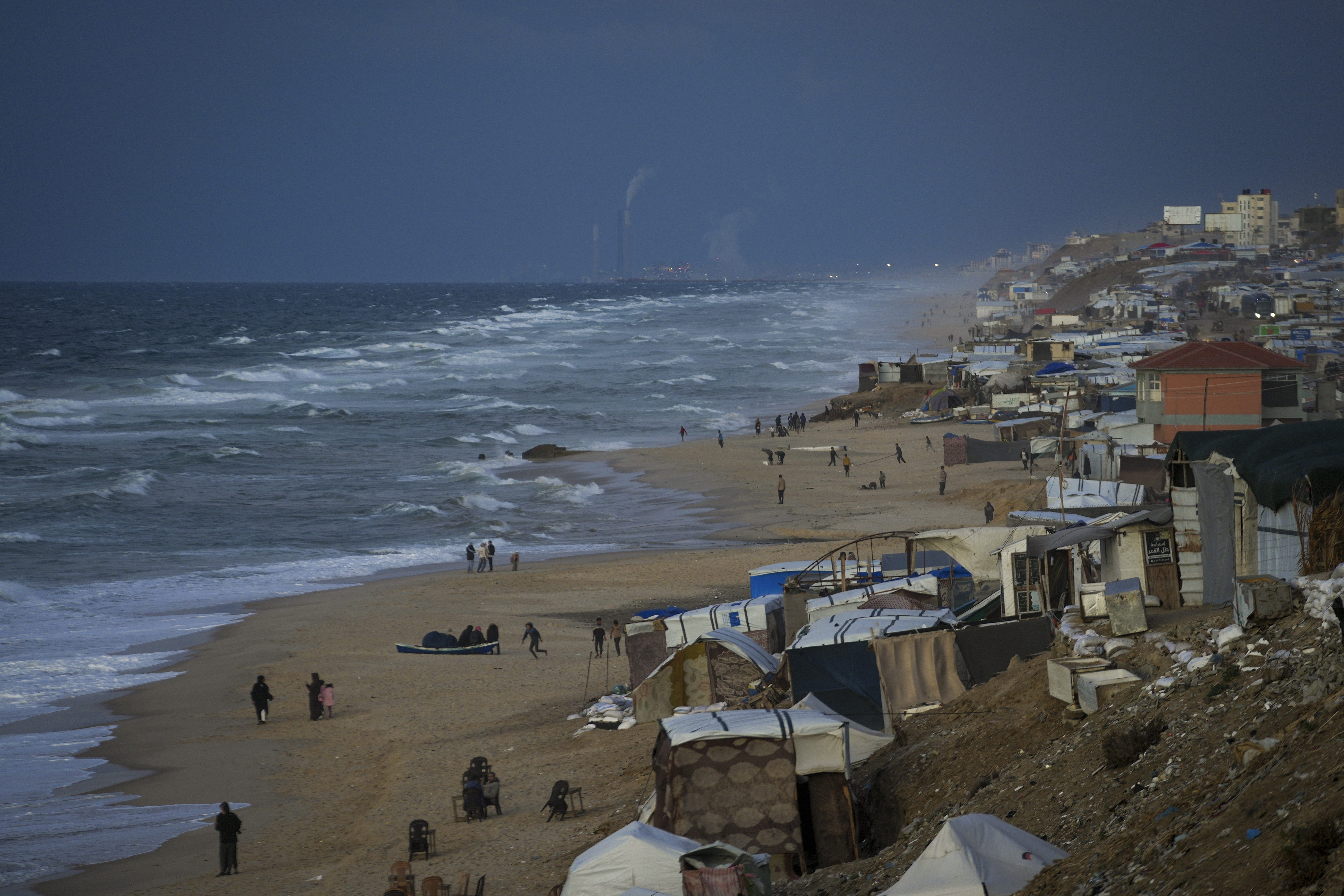 People walk along the beach next to a tent refugee camp for displaced Palestinians in Deir al-Balah, central Gaza Strip, Monday, Dec .30, 2024. (AP Photo/Abdel Kareem Hana)