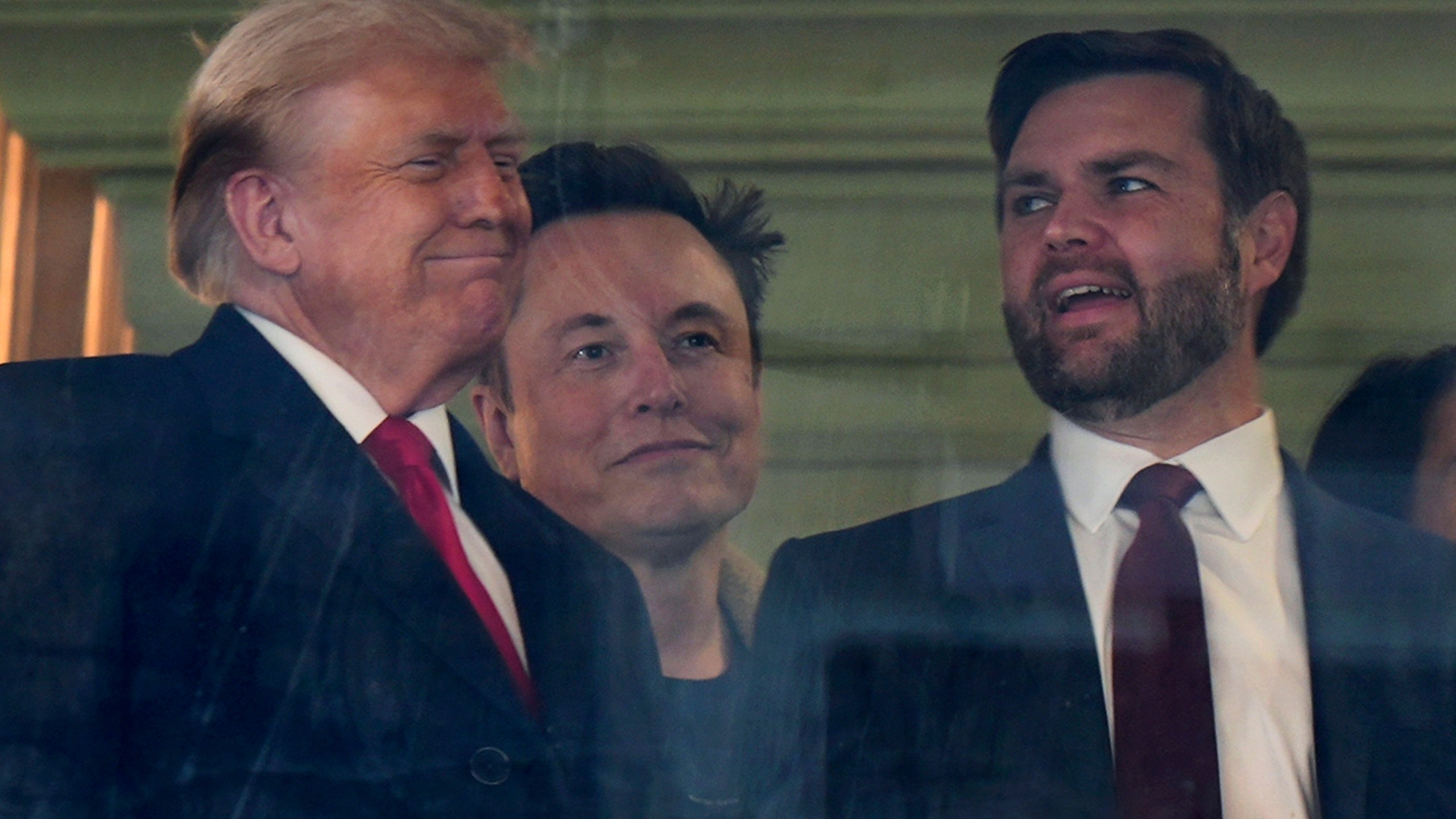 In this picture taken through a window, President-elect Donald Trump, from left, Trump's pick for the planned Department of Government Efficiency Elon Musk and Vice President-elect JD Vance attend the NCAA college football game between Army and Navy at Northwest Stadium in Landover, Md., Saturday, Dec. 14, 2024. (AP Photo/Stephanie Scarbrough)