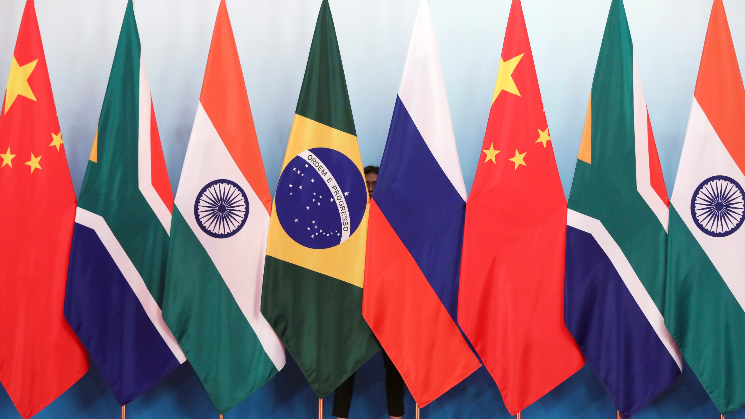 FILE - Staff worker stands behinds national flags of Brazil, Russia, China, South Africa and India to tidy the flags ahead of a group photo during the BRICS Summit at the Xiamen International Conference and Exhibition Center in Xiamen, southeastern China's Fujian Province, Monday, Sept. 4, 2017. (Wu Hong/Pool Photo via AP, File)