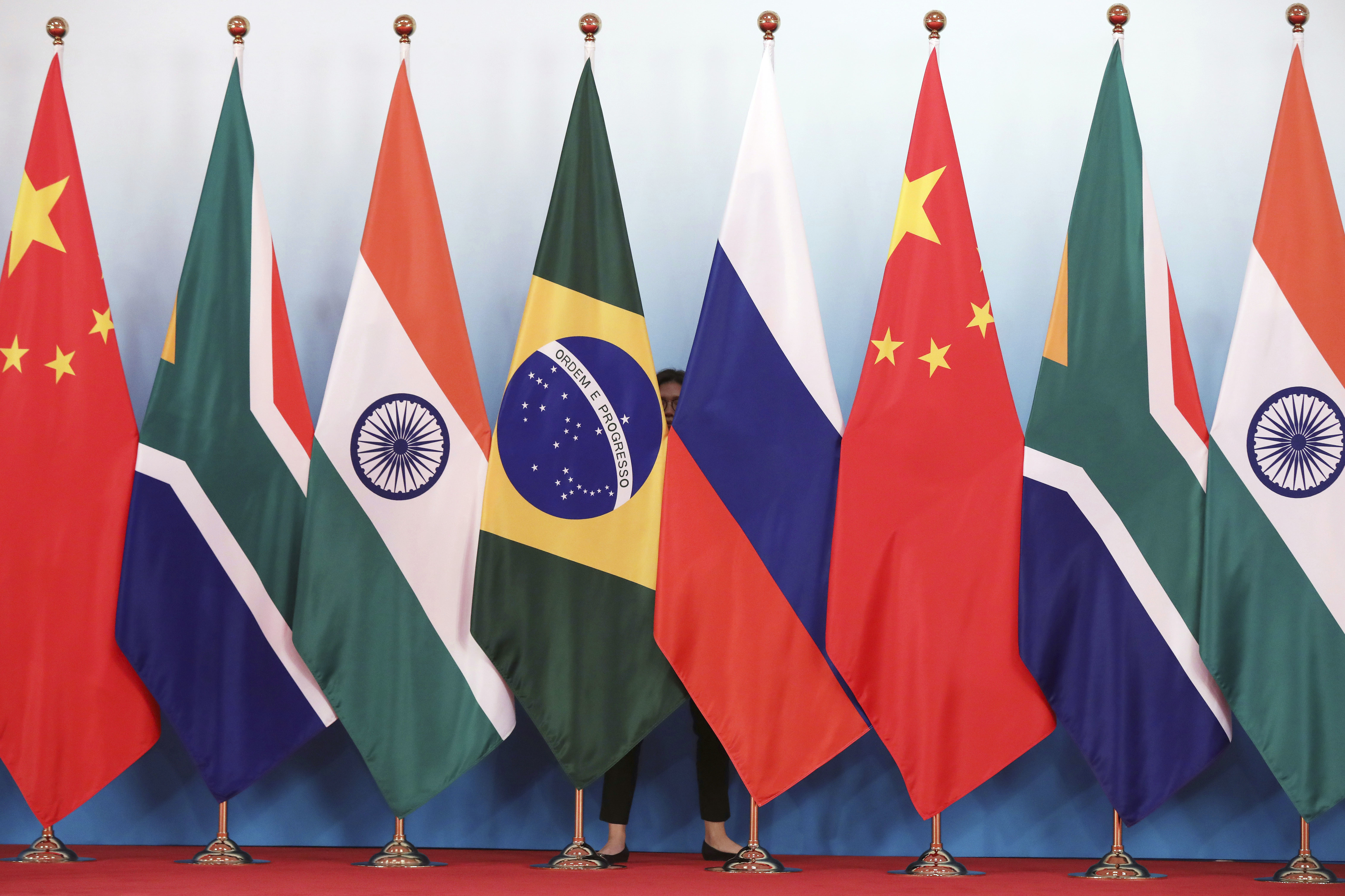 FILE - Staff worker stands behinds national flags of Brazil, Russia, China, South Africa and India to tidy the flags ahead of a group photo during the BRICS Summit at the Xiamen International Conference and Exhibition Center in Xiamen, southeastern China's Fujian Province, Monday, Sept. 4, 2017. (Wu Hong/Pool Photo via AP, File)