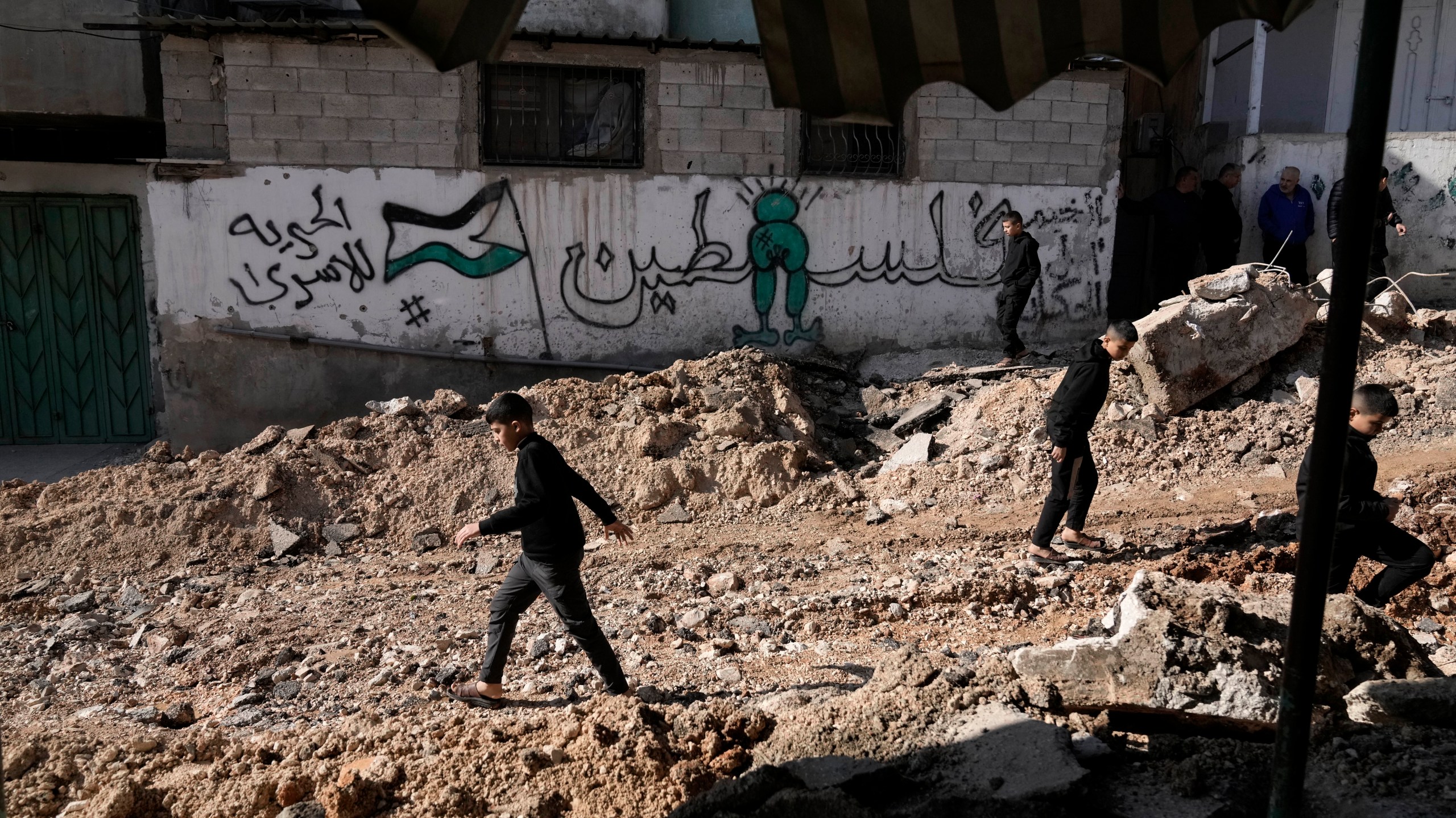 An Arabic mural reading, "Palestine," is the backdrop as Palestinian children walk on a street damaged in Israeli military operation at the Al-Fara'a refugee camp in the West Bank, Tuesday, Jan. 7, 2025. (AP Photo/Majdi Mohammed)